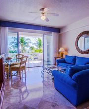 Sala luminosa con sofás azules, espejo redondo y vista al mar desde el balcón con palmeras.