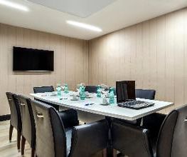 Sala de reunião com mesa retangular, cadeiras pretas e TV na parede.
