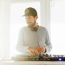 Ein DJ mit Kopfhörern und gestreiftem Shirt mixt Musik an seinem Pult in einem hellen Raum.