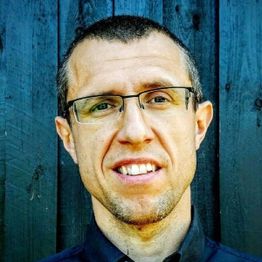 En mand med briller smiler foran en mørk trævæg.