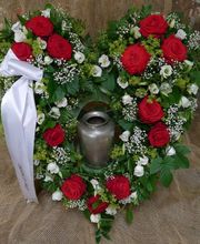 Herzförmiger Blumengesteck mit roten und weißen Rosen um eine Urne auf Juteuntergrund.