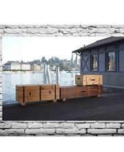 Holzmöbel am Wasser, daneben ein graues Gebäude und im Hintergrund eine Stadtlandschaft.