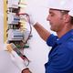 Electrician in blue overalls checks wiring with a multimeter near an electrical panel.