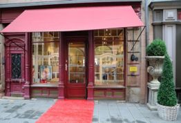 Geschäft mit rotem Vordach und Teppich, große Fensterfront, Pflanze in Pflanzgefäß daneben.