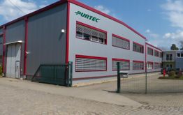 Grau-rotes Fabrikgebäude mit "PURTEC" Logo, gepflasterter Weg und blauer Himmel im Hintergrund.