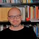 Uomo con occhiali davanti a una libreria piena di libri colorati.