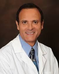 Smiling man in a lab coat and tie against a dark background.