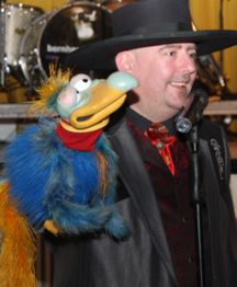 Ein Mann mit Hut und bunter Puppe in der Hand lächelt, Mikrofon im Vordergrund.