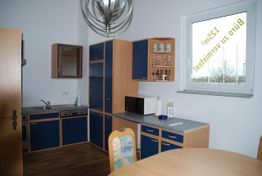 Küche mit blauen Schränken, Mikrowelle und Rundtisch vor Fenster mit "Büro zu vermieten" Zeichen.