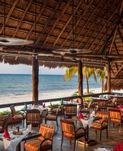 Restaurante junto a la playa con sillas de madera y techo de palma, vista al mar.