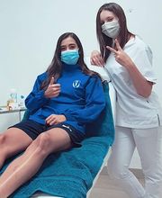 Dos personas con mascarillas, una sentada en una camilla y otra de pie haciendo señal de paz.