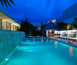 Piscina iluminada de noche, rodeada de palapas y tumbonas, bajo un cielo azul profundo.