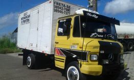 Caminhão amarelo de frete com baú branco, estacionado em área urbana, céu azul ao fundo.