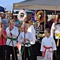 Children in martial arts uniforms hold staffs at an outdoor event with wreaths and people nearby.