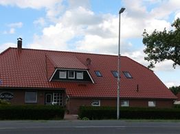 Ziegelgedecktes Haus mit Gaubenfenstern und Laterne, vor einer Hecke unter blauem Himmel.