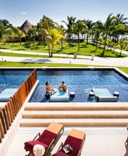 Personas disfrutando en piscina rodeada de palmeras y tumbonas, con jardín y cielo despejado.