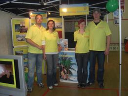 Vier Personen in gelben Hemden stehen an einem Messestand mit Reiseplakaten und Broschüren.
