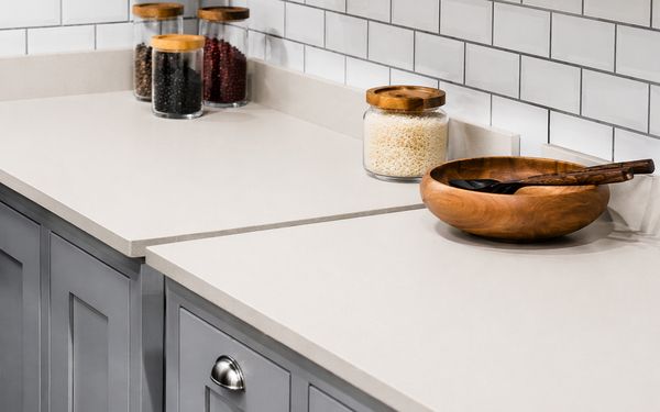 Encimera de cocina de resina en tono beige claro, con superficie lisa, uniforme y sin juntas visibles, acompañada de tarros de vidrio con alimentos y un bol de madera sobre muebles blancos y salpicadero de azulejos.