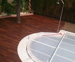 Piscina con cubierta y terraza de madera, rodeada de vegetación y ducha exterior.