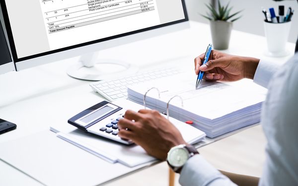 Person sitzt vor Computer, schreibt etwas in Ordner und rechnet mit Taschenrechner