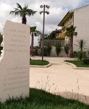 Monumento in un parco con erba, palme ed edifici sullo sfondo, cielo nuvoloso.