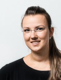 Eine Frau mit Brille lächelt vor weißem Hintergrund. Sie trägt ein schwarzes Oberteil.