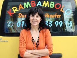 Eine Frau steht lächelnd vor einem gelben Auto mit der Aufschrift "Krambambolo".