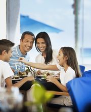 Familia disfrutando de una comida en un restaurante con vista al mar, sonriendo y conversando.
