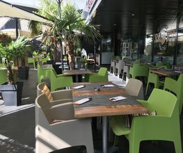 Außenterrasse eines Cafés mit grünen Stühlen, Holztischen und Palmen in Töpfen.
