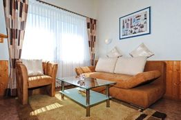 Gemütliches Wohnzimmer mit braunem Sofa, Sessel und Glastisch, eingerahmt von hellen Vorhängen.
