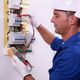 Electrician in blue uniform using a multimeter to check wiring in a circuit breaker panel.