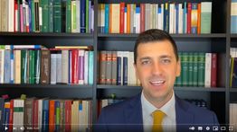 Uomo in giacca e cravatta gialla davanti a una libreria piena di libri colorati.