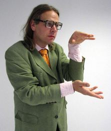 Eine Person im grünen Anzug gestikuliert mit den Händen in einem neutralen Raum.