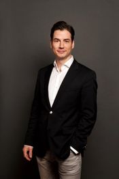 Mann im Anzug vor grauem Hintergrund, lächelnd, mit einer Hand in der Tasche.