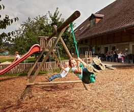 Kinder schaukeln fröhlich auf einem Spielplatz, im Hintergrund ein Haus mit Menschen auf der Terrasse.