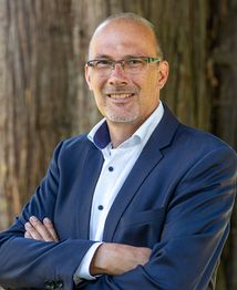 Ich weiß nicht, wer diese Person ist. Mann mit Brille und Anzug lächelt vor einem Baumbereich.