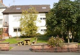 Weißes zweistöckiges Haus mit Garten, Bäumen und Sitzbereich aus Holz, umgeben von Blumenbeeten.