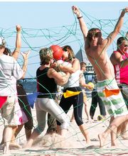 Menschen spielen am Strand ein Spiel mit einem Netz und einem roten Ball unter blauem Himmel.