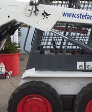 Mini escavatore Bobcat bianco con ruote rosse, accanto a una pianta in vaso rosso e un piccolo gnomo.
