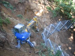Escavatore blu e giallo al lavoro su un pendio, con recinzione metallica instabile.