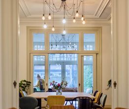 Eetkamertafel met stoelen, bloemenvaas en grote ramen met zicht op persoon en tuin.