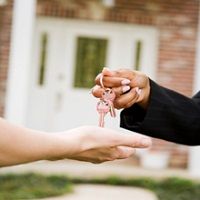 Two hands exchanging keys in front of a house.