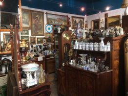 Antiquitätengeschäft mit Gemälden, Möbeln, Gläsern und Keramiken an dekorierten Wänden.