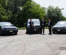 Drei schwarze Mercedes-Benz und zwei Männer in Anzügen auf einem Parkplatz.