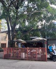 Café ao ar livre com mesas, cercado vermelho e árvores ao fundo. Pessoas conversando.