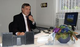 Ein Mann im Anzug telefoniert im Büro. Auf dem Tisch stehen Blumen und Bilderrahmen.