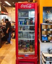Geladeira vermelha de bebidas em loja com garrafas e latas. Pessoa ao fundo, ambiente comercial.