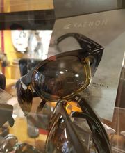 Sonnenbrille auf einem Ausstellungsregal mit einem Kaenon-Poster im Hintergrund.