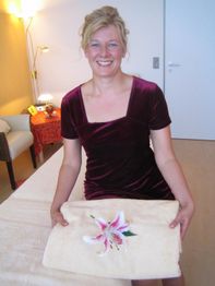 Eine lächelnde Frau in violettem Kleid hält ein besticktes Handtuch mit einer rosa Lilie.