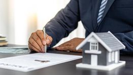 Person signing a document with a model house on the table.